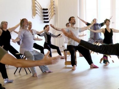 In a large fitness studio a group of people are seated on stools with arms reaching out horizontally from their bodies performing a stretch.